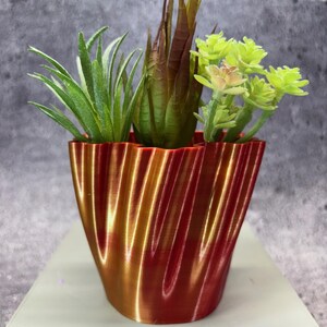 Puede incluir: Una maceta decorativa roja y dorada con un diseño ondulado, llena de suculentas artificiales. El arreglo incluye plantas artificiales verdes, marrones y amarillo-verdosas. La maceta está sobre una superficie gris claro.