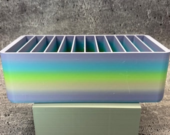 May include: A rectangular organizer with vertical dividers, featuring a gradient of colors from blue to green. The organizer is made of a solid material and sits on a light green surface. The dividers create individual compartments.