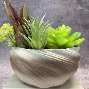 May include: A small, round planter with a swirling, metallic bronze and grey finish containing a variety of artificial succulents. The plants include tall, pointed, reddish-brown leaves, a central spiky green plant, and a rounded green rosette.