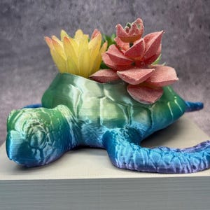 May include: A 3D-printed turtle-shaped planter in shades of blue, green, and purple, holding artificial succulents. The turtle has a textured shell and head, with a yellow and red flower arrangement.