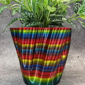 Pode incluir: Um vaso ondulado e listrado de cores, feito de plástico. O vaso está cheio de uma planta verde com manchas brancas.
