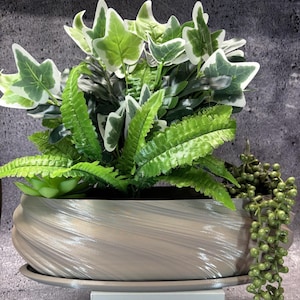 May include: A decorative planter with artificial plants. The light grey planter has a textured, swirling design. It contains green ferns, ivy with white edges, a small succulent, and a trailing plant with small, round green beads. The planter sits on a matching saucer.
