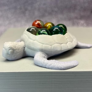 Könnte beinhalten: Eine weiße, 3D-gedruckte Schildkrötenfigur mit bunten Glasmurmeln. Die Schildkröte hat einen strukturierten Panzer und ausgestreckte Flossen. Die Murmeln sind in verschiedenen Farben, darunter Rot, Grün und Gelb. Die Figur liegt auf einer hellen Oberfläche.
