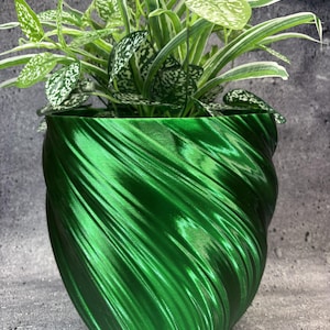 May include: A metallic emerald green plant pot with a spiraling, rippled texture. The pot contains a lush houseplant featuring variegated green and white leaves with speckled and striped patterns, creating a vibrant contrast against the shiny surface.
