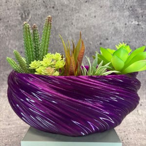 Puede incluir: Una vibrante maceta morada llena de una variedad de suculentas y cactus artificiales. La maceta tiene un diseño texturizado único con un patrón arremolinado. Las plantas artificiales incluyen tonos verdes, amarillos y rojizos, creando una exhibición colorida.