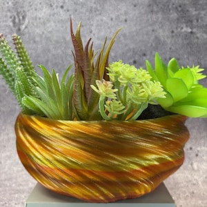 May include: A decorative planter with a swirling, textured design in shades of gold, bronze, and green. The planter is filled with a variety of artificial succulents in green, brown, and yellow-green hues, creating a vibrant and natural aesthetic. The planter is approximately 15 cm wide.