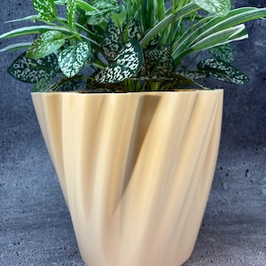 May include: A beige, spiral-shaped planter pot holding a green plant with white-speckled leaves. The pot has a unique, twisted design, and the plant's leaves are a mix of green and white. The pot is sitting on a gray surface.