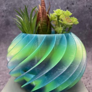 May include: A spiral-shaped planter in shades of blue and green, holding a variety of small, artificial succulents. The planter's design features a series of curved, overlapping segments, creating a dynamic, textured appearance. The plants include green and brown faux succulents.