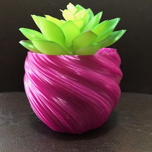 May include: A magenta-coloured pot with a spiral texture contains a bright green and yellow artificial succulent. The plant pot has a unique, swirling design. The succulent has multiple layers of green leaves with yellow tips.