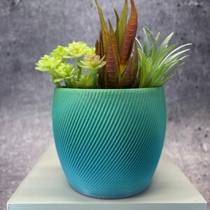 May include: A small, teal-colored planter with a spiral design, filled with a variety of artificial succulents. The succulents include green, yellow, and reddish-brown plants. The planter sits on a light gray surface, creating a modern, decorative accent.