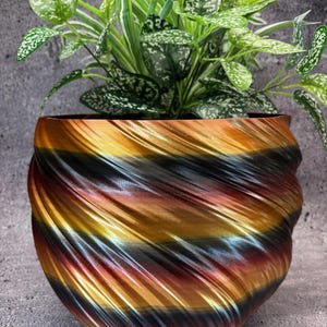 May include: A decorative planter with a swirling pattern in shades of gold, black, red, and blue. The planter is made of a smooth, shiny material and has a rounded shape.