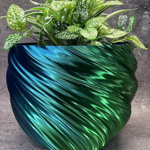 May include: A decorative planter with a swirling design in shades of blue and green, holding a vibrant green plant with white-speckled leaves. The planter has a glossy finish and sits on a textured grey surface.