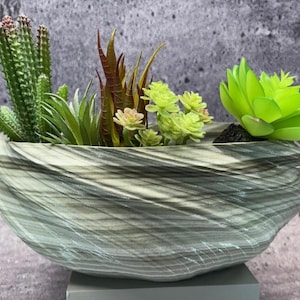 Może przedstawiać: Dekoracyjna, szaro-biała, ceramiczna donica w paski wypełniona różnorodnymi sztucznymi sukulentami i kaktusami. Kompozycja zawiera zielone, czerwone i żółto-zielone rośliny, tworząc naturalną estetykę. Donica ma owalny kształt.