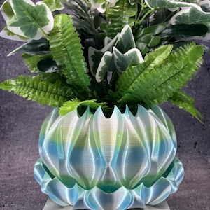 May include: A 3D-printed planter with a scalloped, petal-like design featuring a gradient of iridescent blue, green, and white colors. The planter holds a lush arrangement of artificial green and white variegated ivy and fern-like foliage.