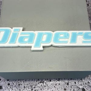 Puede incluir: Un letrero blanco y azul claro con la palabra "Diapers", con relleno azul claro y contorno blanco, descansa sobre una superficie gris. El letrero es un diseño estilizado en tres dimensiones, con la palabra "Diapers" en negrita, sin serif.