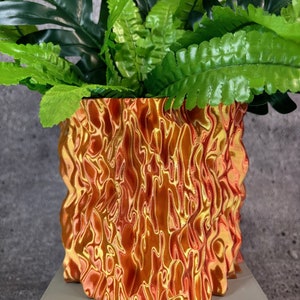 May include: A decorative plant pot with a textured, flame-like design in shades of orange and gold. The pot is filled with vibrant green artificial plants, including large, tropical-style leaves and smaller fern-like foliage. The pot sits on a light gray surface.
