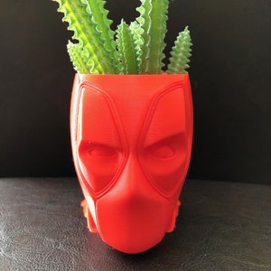 Puede incluir: Un macetero rojo con forma de cabeza de Deadpool con una planta suculenta en su interior.