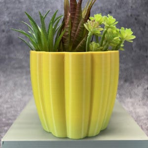 May include: A yellow, ribbed planter pot filled with assorted faux succulents. The artificial plants include green, brown, and light green foliage. The pot is sitting on a light green surface against a grey background.