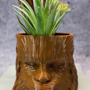 May include: A brown, 3D-printed planter shaped like a character's face, holding a variety of faux succulents and greenery. The planter has a textured surface and detailed facial features. The plants include green and yellow succulents and brown and green foliage.