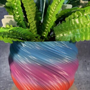 May include: A colourful, swirl-patterned planter with a gradient of blue, pink and orange. The planter is filled with green leafy plants.