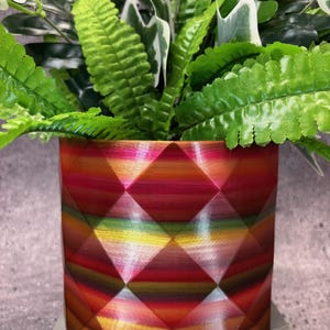 Può includere: Un vaso da fiori geometrico con un motivo a strisce multicolore in tonalità di rosso, arancio, giallo e verde. Il vaso è realizzato in un materiale liscio e lucido e ha una forma esagonale.