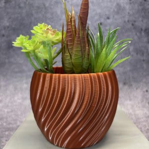 May include: A small, brown, decorative planter with a textured, swirling design, filled with a variety of artificial succulents in shades of green and brown. The planter sits on a white surface against a gray background.