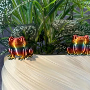 May include: Two small, colorful frog figurines sit on the edge of a white, textured planter. The frogs have a rainbow metallic finish, with shades of red, orange, yellow, and green. Green leafy plants fill the background.