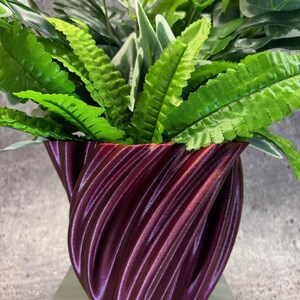 Op de afbeelding: Een bordeauxrode, 3D-geprinte plantenpot met een wervelend ontwerp, gevuld met levendige groene kunstplanten. De pot heeft een textuuroppervlak en staat op een witte plank. De planten omvatten varens en andere bladvariëteiten.