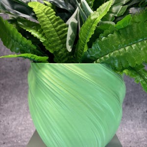 May include: A green plant pot with a textured, swirling design, filled with green foliage. The leaves are a mix of dark and light green. The pot is on a light grey surface. The pot's design adds a modern touch to the greenery.