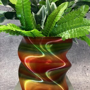 May include: A decorative vase with a swirling design in shades of red, green, and gold, holding an arrangement of artificial green and white-veined leaves. The vase has a unique, textured appearance.
