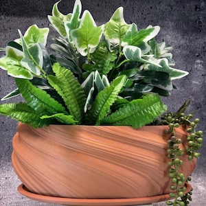 Op de afbeelding: Een terracotta-kleurige, decoratieve plantenbak met een wervelend ontwerp, gevuld met een verscheidenheid aan groene en witte kunstplanten. De opstelling omvat varens, klimop en hangplanten, waardoor een weelderige uitstraling ontstaat. De plantenbak staat op een bijpassende schotel.