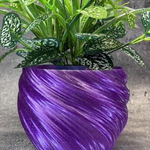 Könnte beinhalten: Ein dekorativer, glitzernder, violetter Blumentopf mit einem gewellten Design. Er enthält eine grüne Pflanze mit weiß gefleckten Blättern. Die Oberfläche des Topfes reflektiert das Licht. Ein stilvolles Element für die Inneneinrichtung, für verschiedene Pflanzen.