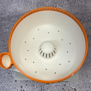 May include: A round, white plastic colander with a bright orange rim and a small, circular side handle. The interior features a central raised dome with vertical slits and a series of small drainage holes distributed across the base.