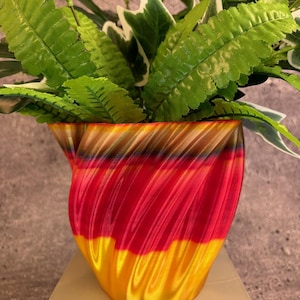 Op de afbeelding: Een rode, gele en gouden swirl-pot met een groene, bladerrijke plant erin. De pot is gemaakt van een glanzend, glad materiaal.