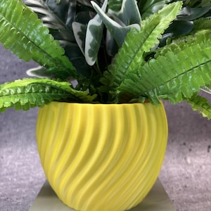 May include: A bright yellow, spherical planter with a textured, spiraling vertical ribbed pattern. The planter contains a dense arrangement of artificial green foliage, including serrated fern-like leaves and broad, dark green leaves with white edges.