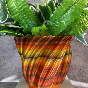 May include: A decorative, orange, yellow, and brown striped vase holds green and white leafy plants. The vase has a unique, swirling design and sits on a white surface. The plants add a natural element to the decorative vase.
