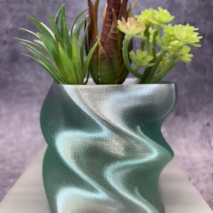 May include: An iridescent green and silver planter with a twisted design, containing artificial succulents. The faux plants feature green, brown, and yellow-green foliage. The planter is displayed on a light-coloured surface. The planter's dimensions are approximately 10 cm.