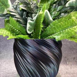 May include: A dark, iridescent black planter with a spiraling, ribbed texture containing a lush arrangement of artificial green and white variegated leaves and fern-like foliage, creating a contrast between the dark container and bright plants.
