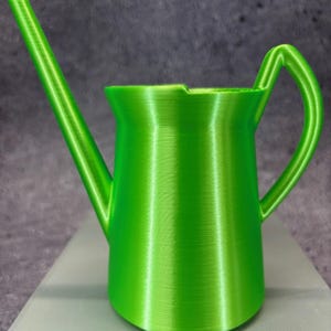 Puede incluir: Una regadera de plástico verde brillante con un pico largo y un asa. La regadera tiene una superficie lisa y texturizada.