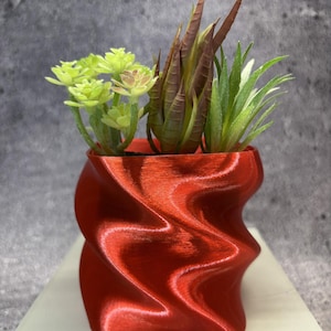 Puede incluir: Una maceta roja metálica impresa en 3D con una silueta retorcida y ondulada. El recipiente contiene tres plantas suculentas artificiales distintas: una con hojas redondeadas de color verde claro, una con hojas puntiagudas marrones y una con follaje verde espinoso.