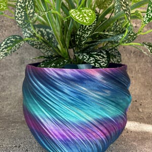 May include: A colourful, swirled planter in shades of blue, teal, and purple holds a lush green plant with speckled leaves. The planter's design features a textured, flowing pattern, creating a dynamic aesthetic. A decorative piece for home decor.