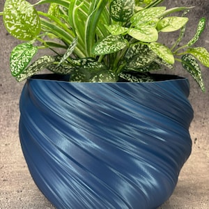 Puede incluir: Una maceta azul texturizada en forma de espiral llena de plantas de hojas verdes y blancas. La maceta tiene un diseño único en espiral y está hecha de un material azul oscuro sólido. Las plantas tienen una variedad de tonos verdes y motas blancas.
