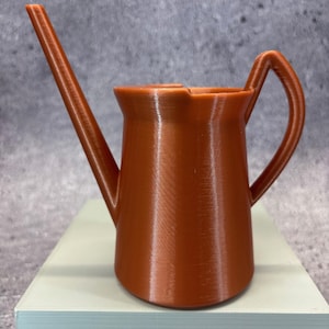 Könnte beinhalten: Eine kleine, terrakottafarbene Gießkanne mit einem langen, schlanken Ausguss und einem gebogenen Griff. Die Gießkanne besteht aus einem festen Material und steht auf einer hellblauen Oberfläche. Das Design ist einfach und funktional.