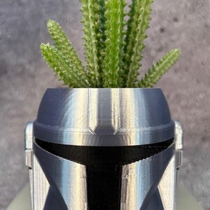 May include: A 3D-printed planter shaped like a Mandalorian helmet, in shades of gray and black, holding a green cactus. The planter has detailed lines and a smooth finish. A unique decorative item for plant lovers and Star Wars fans.