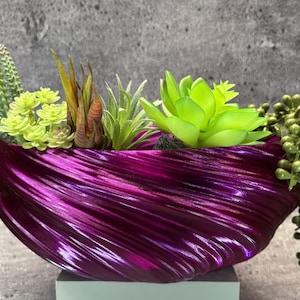 May include: A deep magenta, oval-shaped planter with a textured, swirling ribbed surface. The planter is filled with an assortment of artificial succulents, including tall green spiky plants, light green rosettes, and a trailing string of pearls.
