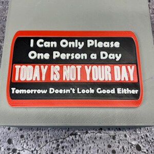May include: A rectangular sign with a black background and red border. The text "I Can Only Please One Person a Day" is in white. Below, in red and white, it reads "TODAY IS NOT YOUR DAY." The bottom line says "Tomorrow Doesn't Look Good Either."