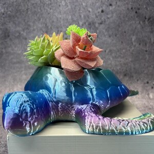 May include: A 3D-printed turtle-shaped planter in shades of blue, purple, and green, holding a variety of artificial succulents. The turtle has a textured shell and is resting on a white surface.