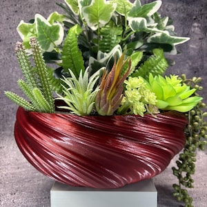 May include: A deep red, spiraled, bowl-shaped planter filled with an assortment of artificial greenery, including variegated ivy, tall green cacti, spiky air plants, light green succulents, and a trailing string of pearls plant.