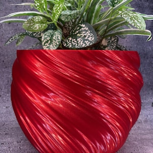 Peut inclure: Un pot de plante rouge vif, texturé avec un motif tourbillonnant, contenant une plante verte luxuriante avec des feuilles tachetées de blanc. Le pot est fait d'un matériau brillant et repose sur une surface grise. La plante a des feuilles vertes avec des taches blanches.