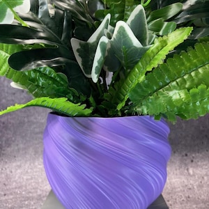 May include: A vibrant purple planter with a swirling, textured exterior design holds a dense arrangement of artificial greenery, including dark green fern fronds and broad, variegated leaves with white edges, creating a lush, layered botanical display.